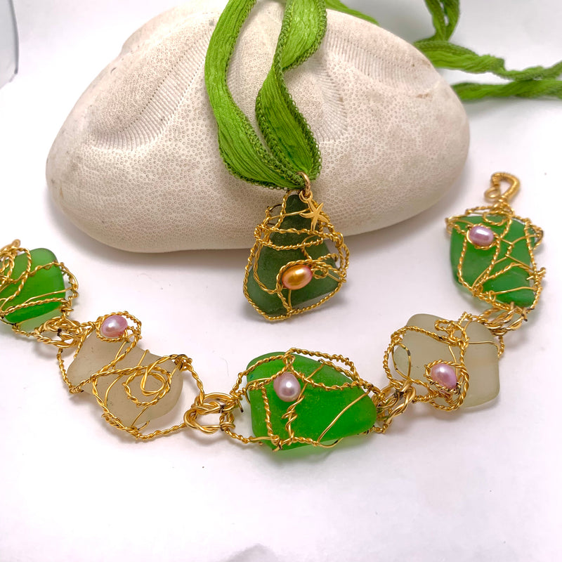 A green and clear sea glass jewelry set with gold wire wraps and small pearls, featuring a pendant on a green ribbon and a matching bracelet, displayed on a white stone.