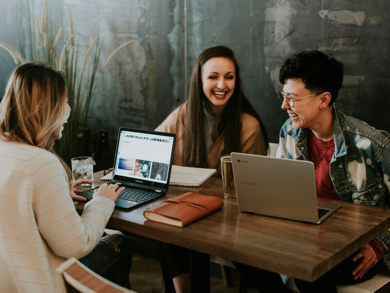 Three people sit at a wooden table with laptops, notebooks, and drinks, smiling and talking together. The background is a dark, textured wall with a plant in the corner.