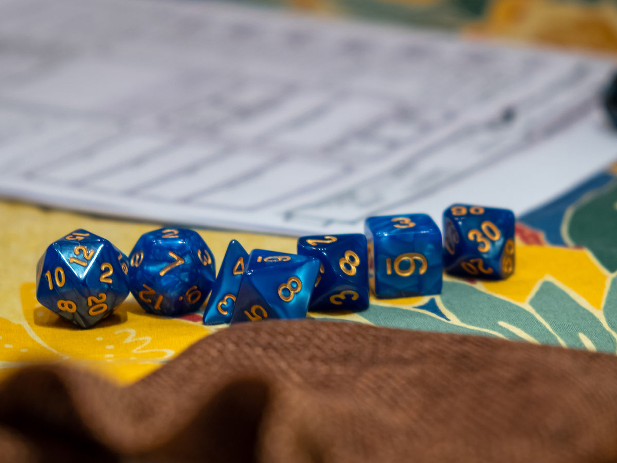 A set of seven blue polyhedral dice with gold numbers sits on a colorful surface, with a blurred character sheet and fabric in the background.
