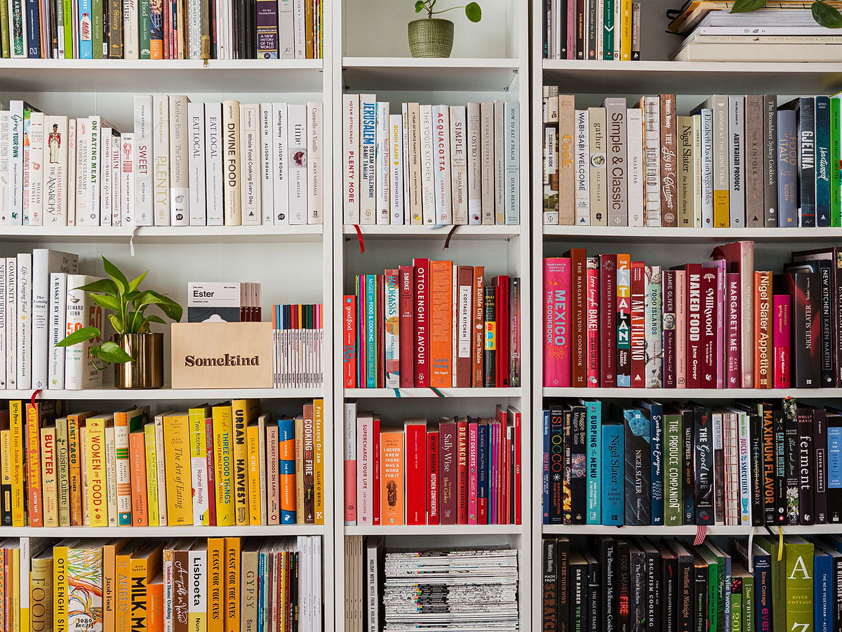 A bookshelf filled with books organized by color, creating a gradient from white and beige on the left, to red, yellow, and blue, to black on the right. Two small green plants and a brown bag are also visible on the shelves.