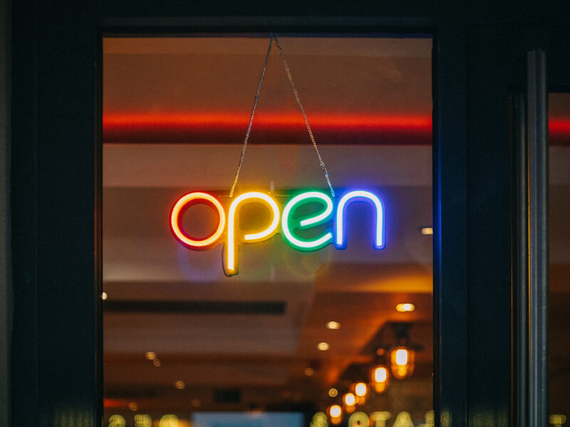 A neon sign with the word "open" in rainbow colors hangs in a window. The background shows a warmly lit interior with ceiling lights.