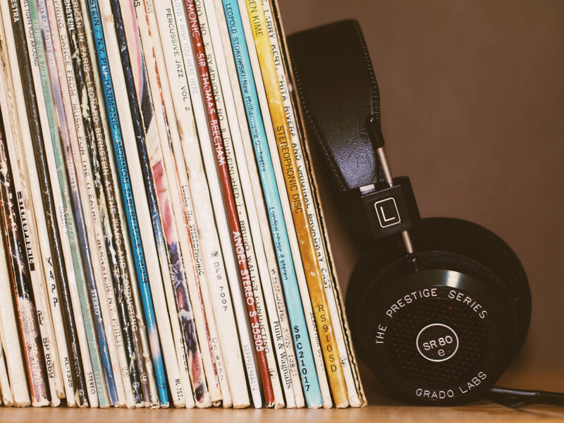 A row of vinyl records stands upright next to a pair of black headphones labeled “SR80” from Grado Labs’ Prestige Series, with the headphones resting against the records. The background is plain and neutral-colored.