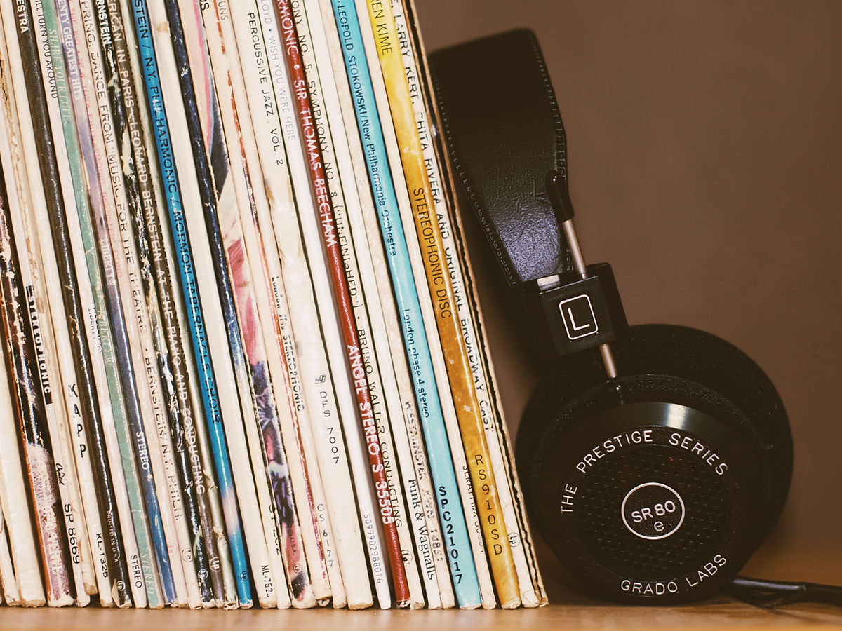 A row of vinyl records stands upright next to a pair of black headphones labeled “SR80” from Grado Labs’ Prestige Series, with the headphones resting against the records. The background is plain and neutral-colored.