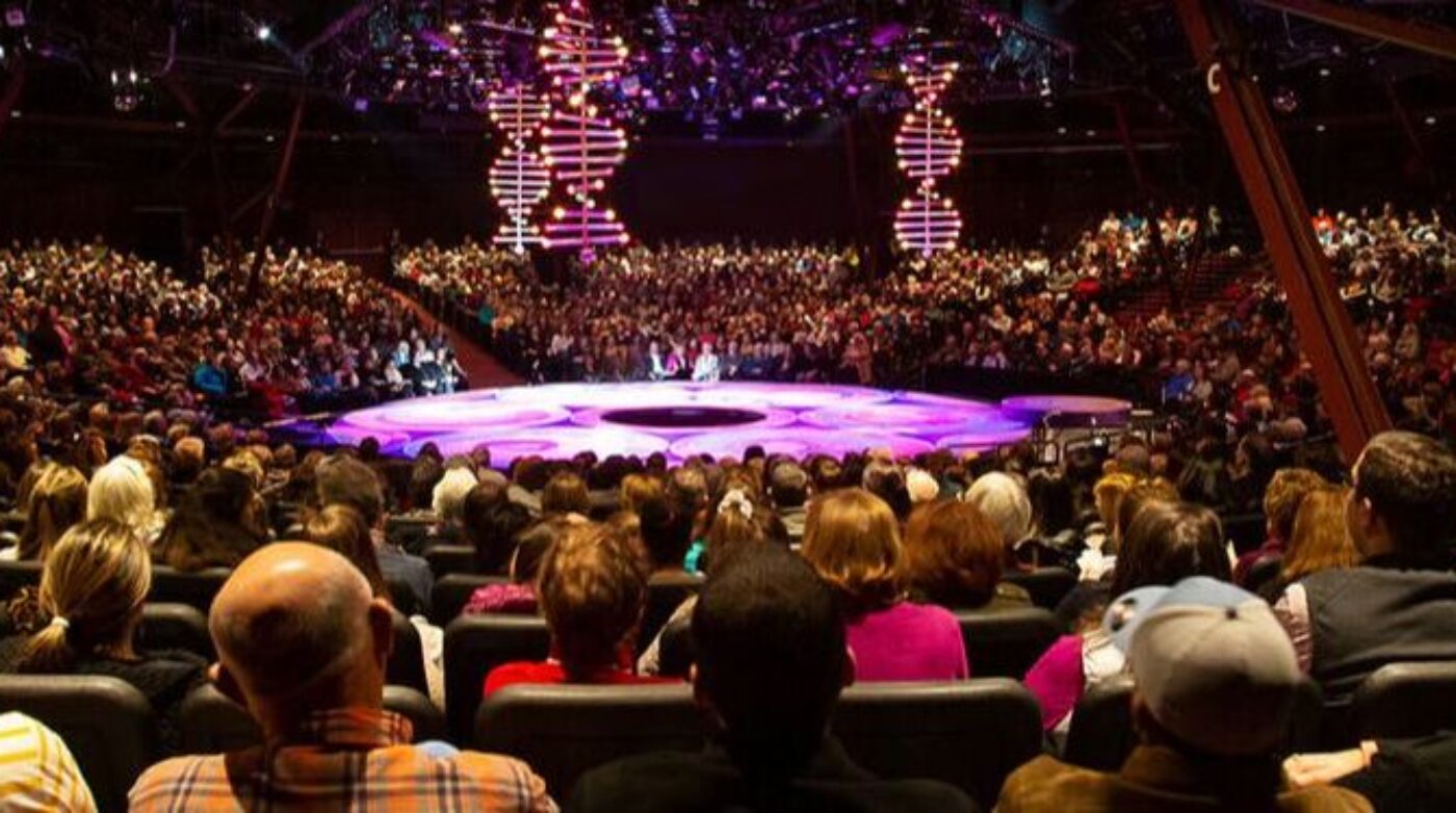 A large audience is seated in an auditorium facing a circular stage with purple lighting. Above the stage, illuminated structures resembling DNA helixes set the scene for Rock of Ages at North Shore Music Theatre. The venue is filled with people.