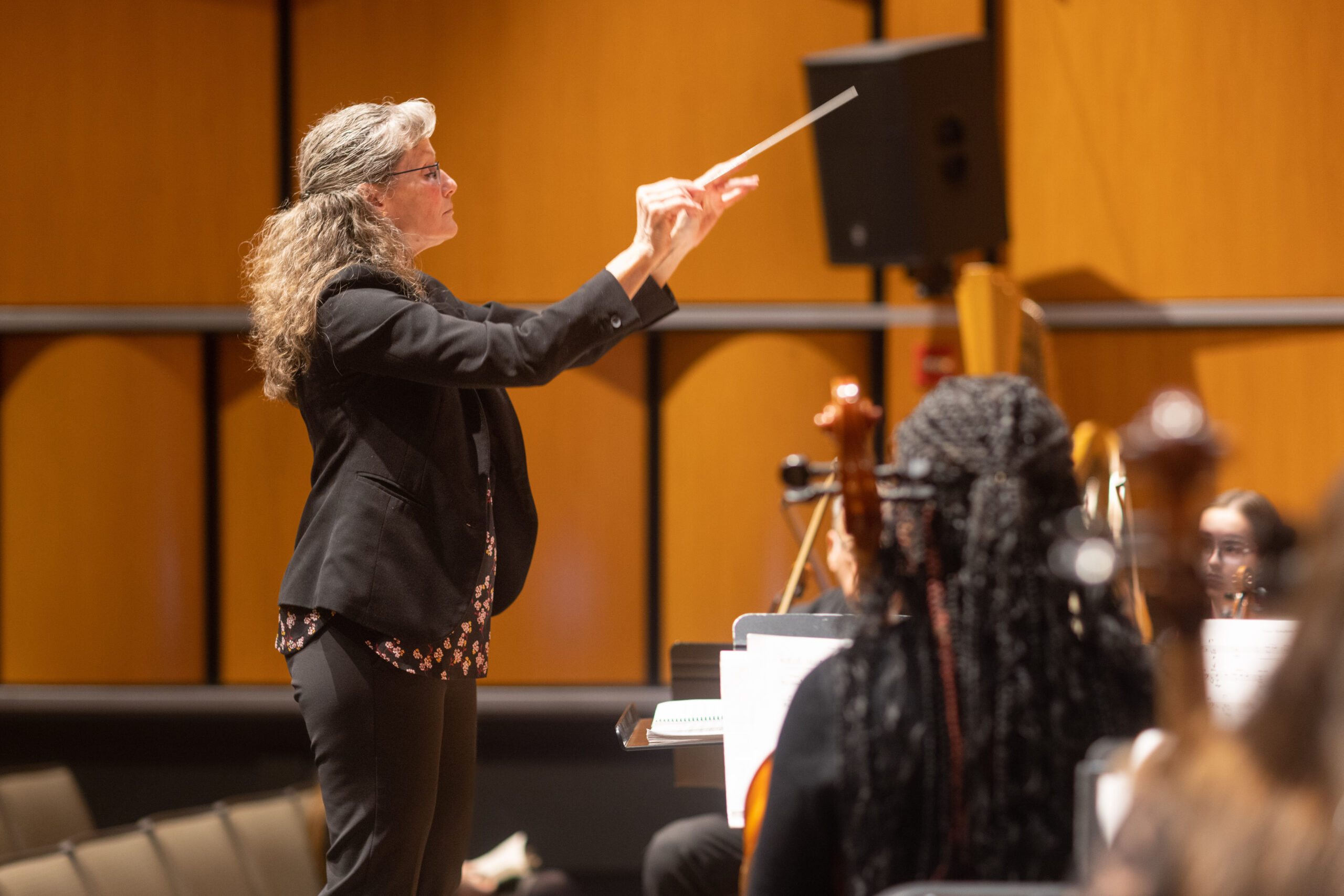 At the Salem State Summer Orchestra concert, a woman in a black suit conducts an orchestra, musicians seated before her. Wooden panels and a speaker line the background of this elegant concert hall scene.