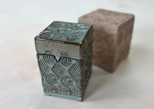 Two rectangular ceramic boxes with lids are shown. The front box is a blue-green, glossy Textured Keepsake Box with geometric patterns; the beige box behind is matte with a similar design. Both rest on a light, smooth surface.