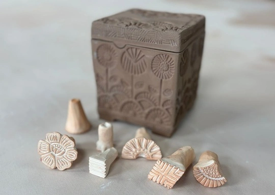 A square clay box with floral patterns stands behind seven small custom stamps, each featuring different carved designs, including flowers and geometric shapes. The hand-built structures rest on a light-colored surface.
