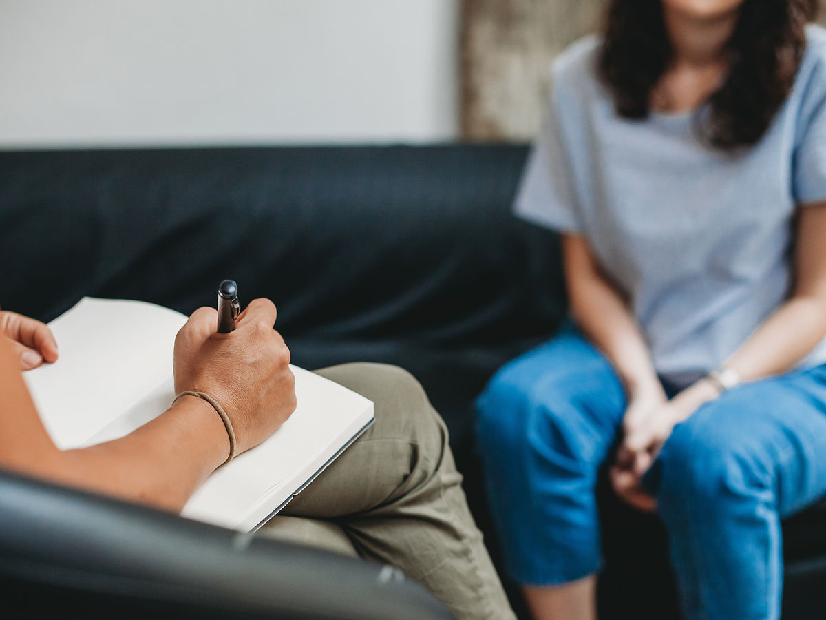 Two people sit indoors. One person writes in a notebook, while the other sits across from them with hands clasped. Both are partially visible; the focus is on the writing hand and the notebook. The setting suggests a conversation or counseling session.