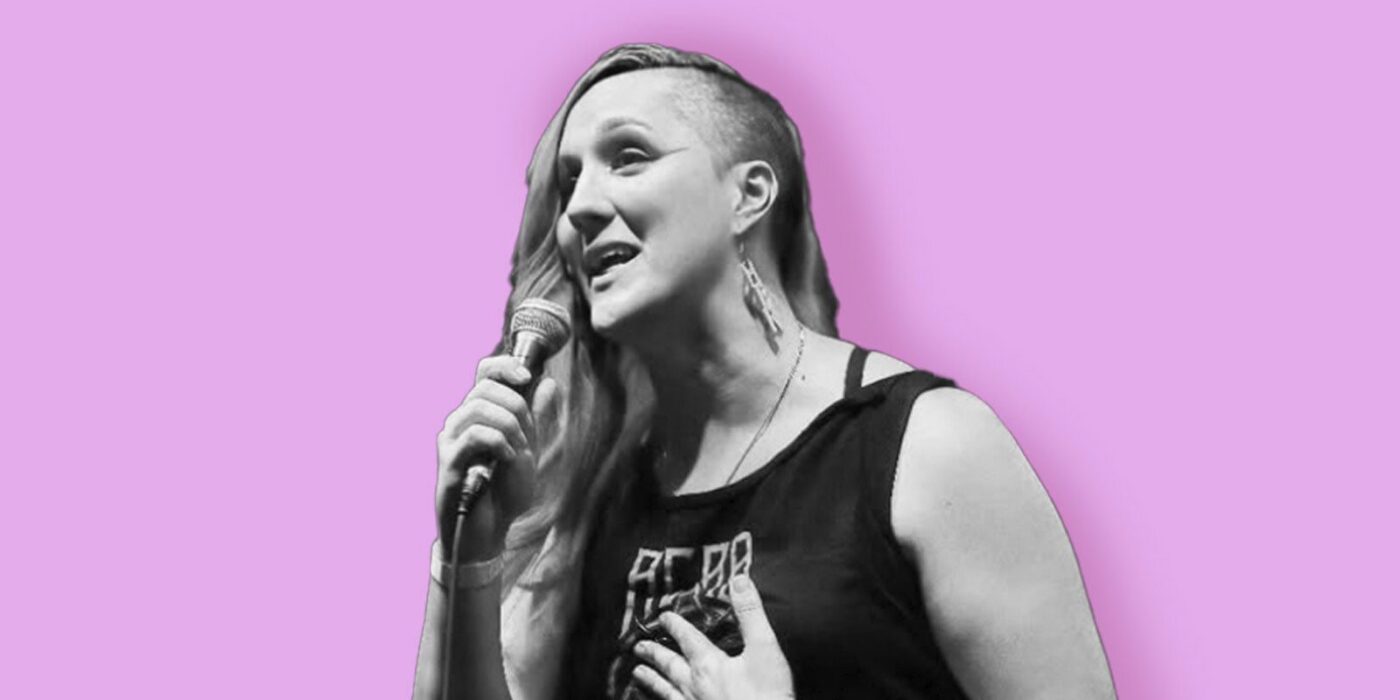 A person with long hair on one side and a shaved side holds a microphone and speaks or sings. They wear a sleeveless top and earrings. The background is a solid light purple color. The image is mostly black and white except for the background.