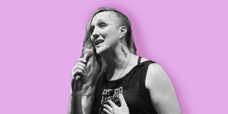 A person with long hair on one side and a shaved side holds a microphone and speaks or sings. They wear a sleeveless top and earrings. The background is a solid light purple color. The image is mostly black and white except for the background.