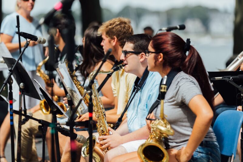 A group of young musicians sit outdoors in a row, holding saxophones and reading sheet music. Microphones are set up nearby, and other people and trees are visible in the background. The setting appears to be a rehearsal or performance.