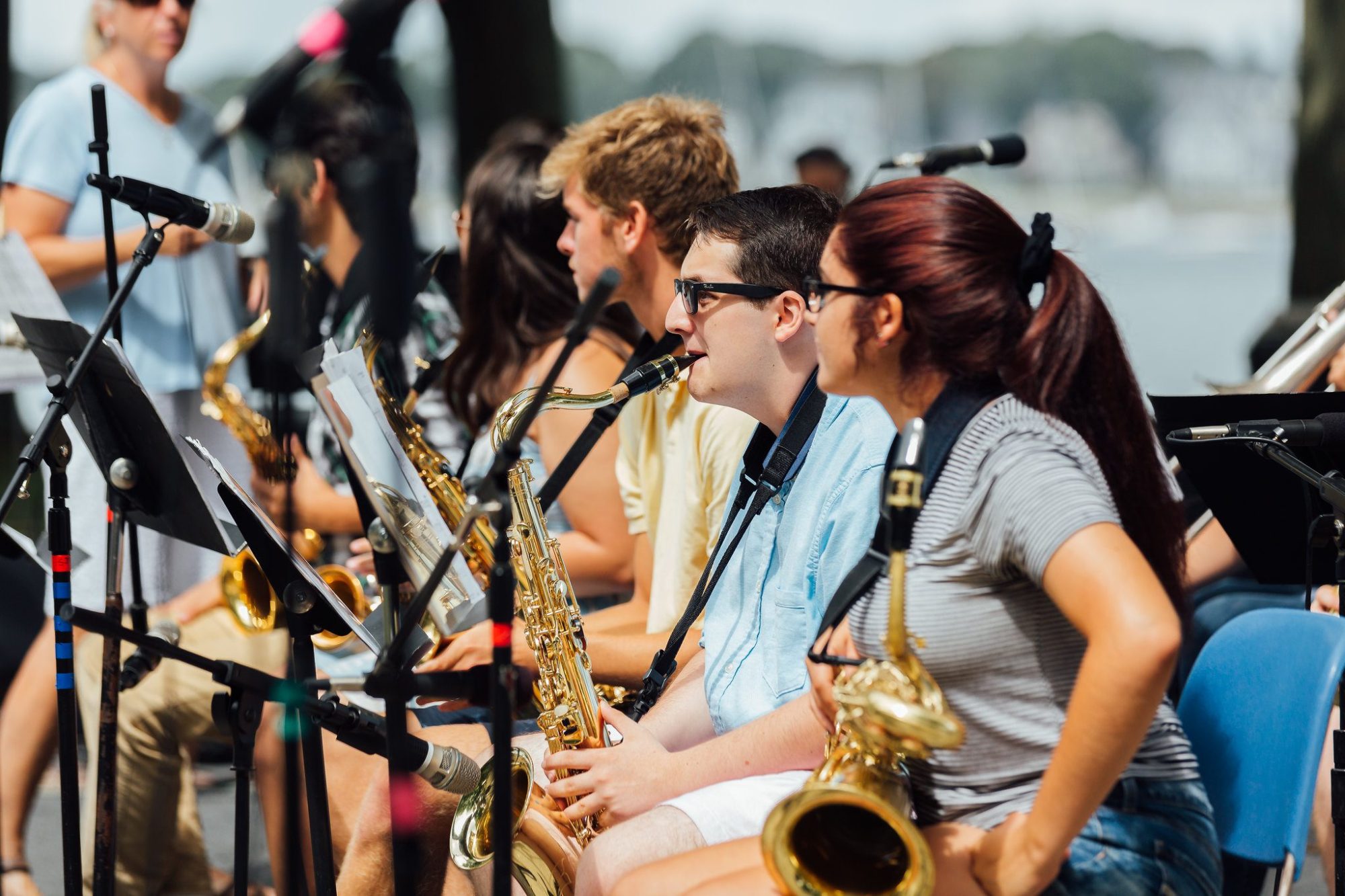 A group of young musicians sit outdoors in a row, holding saxophones and reading sheet music. Microphones are set up nearby, and other people and trees are visible in the background. The setting appears to be a rehearsal or performance.