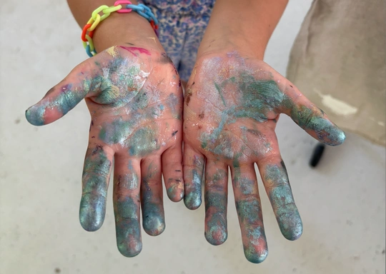 A child, hands coated in blue and green paint, stands on a neutral surface. A colorful chain bracelet hugs one wrist—capturing a moment of magical creations at Salt Studio.