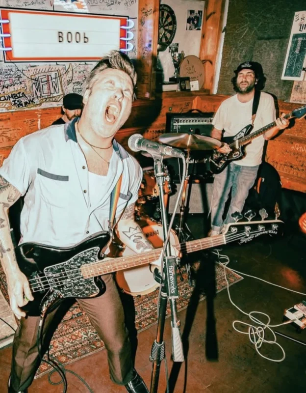 A musician sings passionately into a microphone while playing bass guitar. Another band member in the background plays electric guitar. The scene takes place in a small venue with posters and writing on the walls, and an illuminated sign overhead.