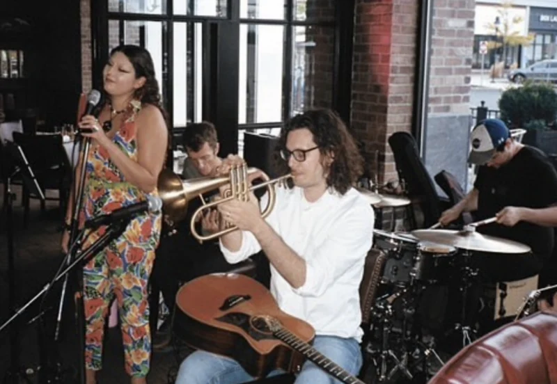A woman sings into a microphone beside three male musicians playing trumpet, drums, and guitar inside a brick-walled venue with large windows.