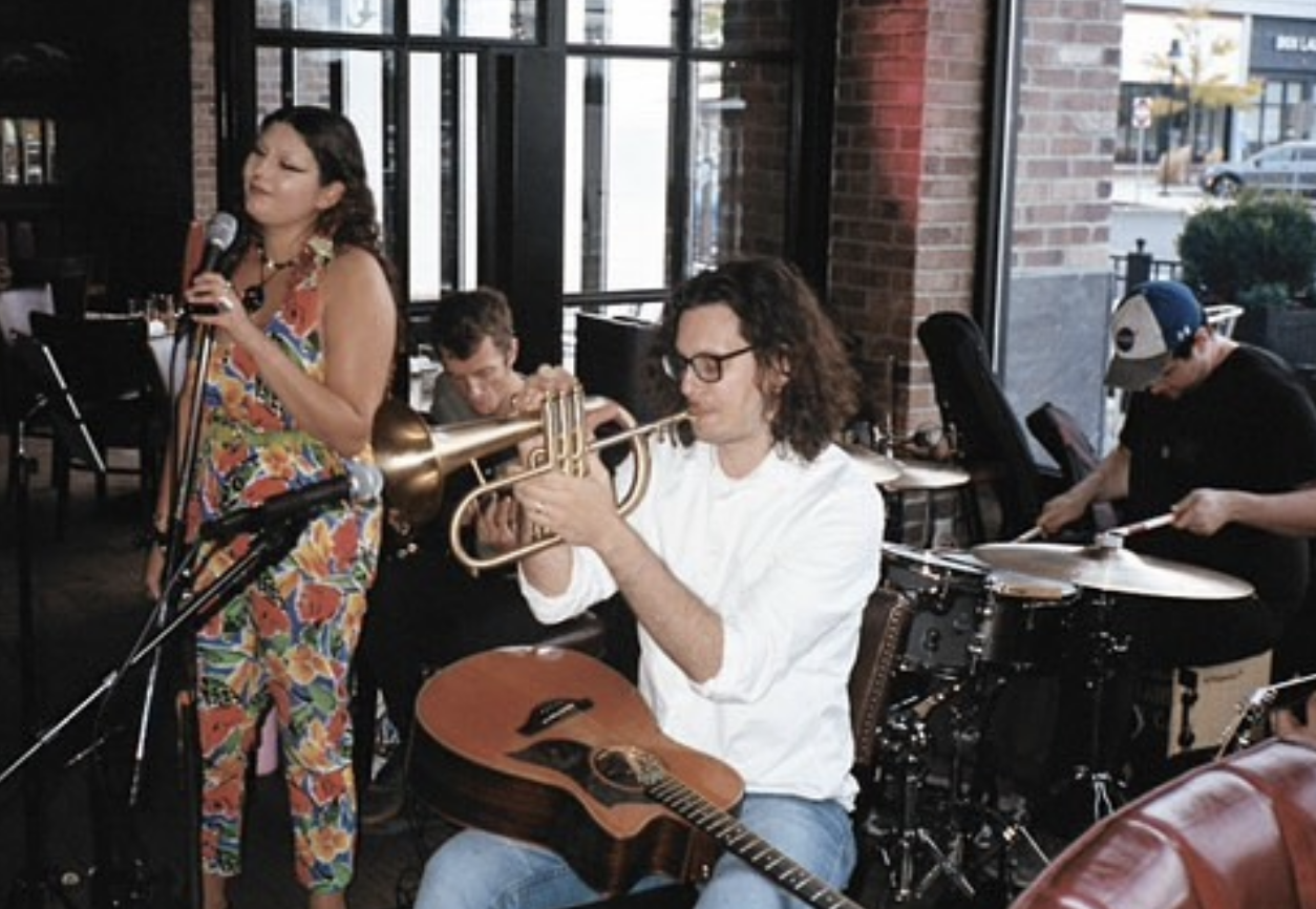 A woman sings into a microphone beside three male musicians playing trumpet, drums, and guitar inside a brick-walled venue with large windows.