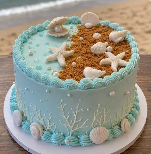 A round cake with pale blue icing, decorated with white chocolate seashells, starfish, seaglass-inspired accents, brown crumbs resembling sand, and piped waves and coral. The background shows a beach and ocean.
