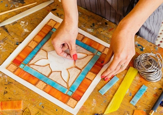 A person arranges colored glass pieces on a geometric pattern for a stained glass, or vitrales, project, using their hands to position the pieces on a wooden work table. Various glass strips and tools are also visible nearby.