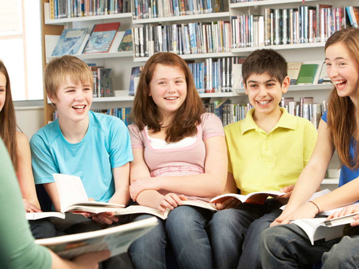Five teenagers sit in a row in a library, each holding an open book. They are smiling and laughing while facing an unseen person across from them. Shelves filled with books are visible in the background.