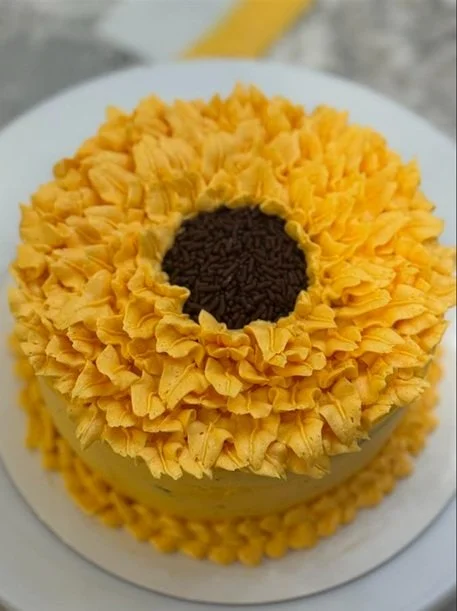 A round cake decorated to look like a sunflower with yellow frosting petals and chocolate sprinkles in the center sits on a white plate.