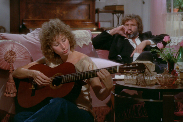 A woman with curly hair sits on a sofa playing an acoustic guitar, while a man in a suit sits in the background smoking and watching her. The room is decorated with pink cushions and a vase with a rose.