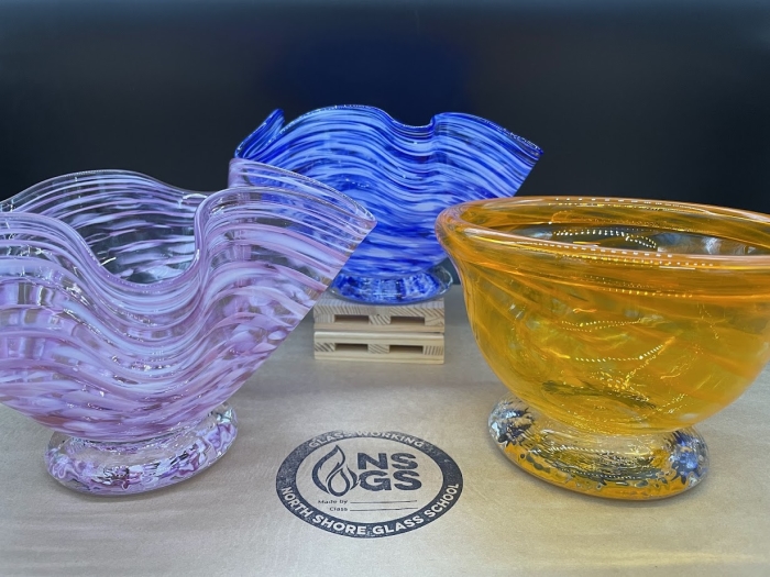 Three wavy glass bowls in purple, blue, and orange are displayed on a table. The blue bowl sits on a small wooden stand. A round logo for North Shore Glass School is visible on the tabletop in front of the bowls.