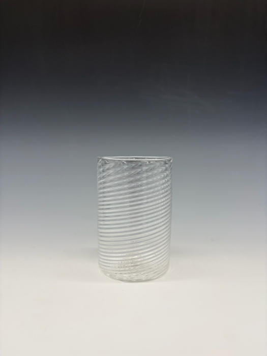 A clear, cylindrical glass tumbler with diagonal ribbed detailing sits upright on a plain, light surface. The background fades from dark gray at the top to white at the bottom, creating a simple, gradient backdrop.