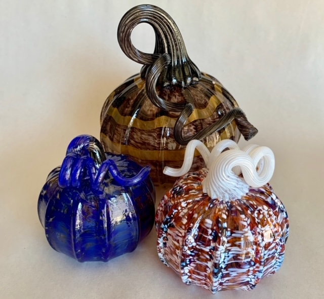 Three decorative glass pumpkins in blue, brown striped, and orange with white swirls are arranged together on a neutral background. Each pumpkin has a curled glass stem in a different color.