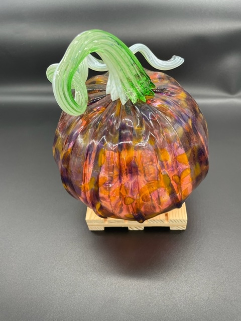 A decorative glass pumpkin with a green curly stem sits on a small wooden pallet against a plain dark background. The pumpkin features orange, purple, and yellow hues with a glossy finish.