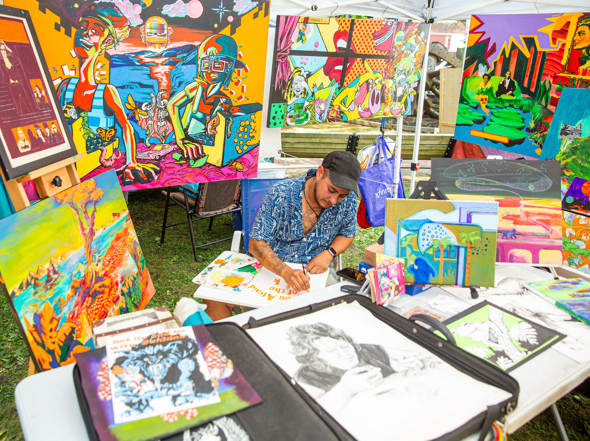 An artist sits at a table drawing, surrounded by vivid paintings and sketches under a white tent at an outdoor art fair—one of the unique things to do in Downtown Lynn. Art supplies and black-and-white sketches are displayed on the table.