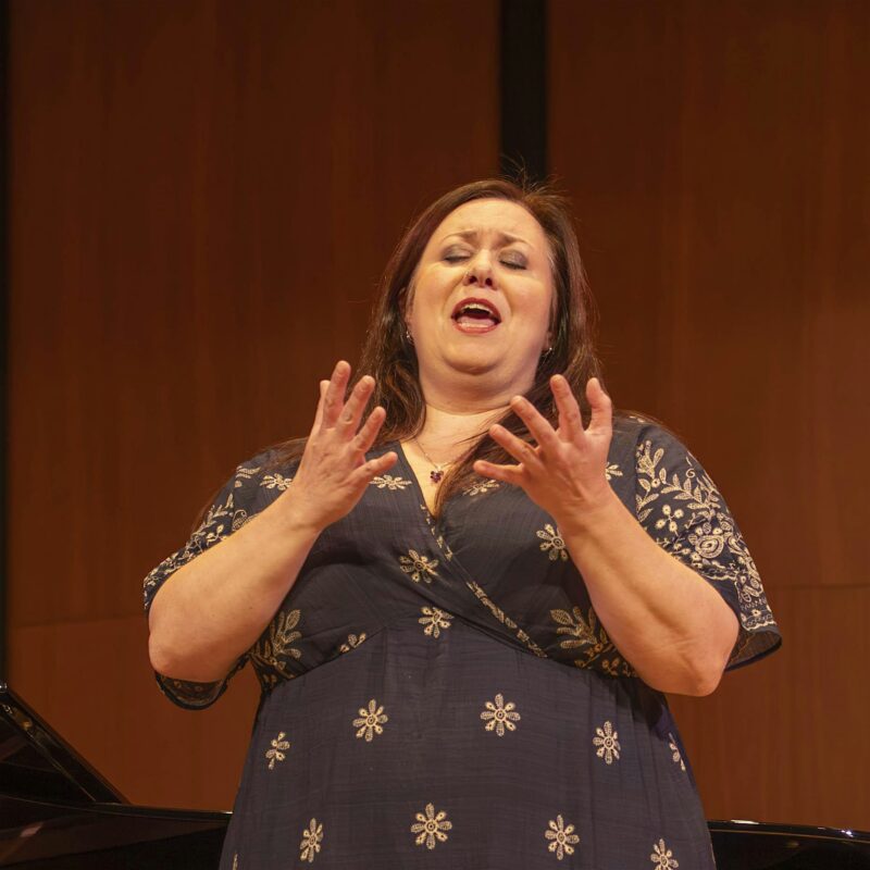 A woman in a dark dress with floral patterns stands on stage, singing with her eyes closed and hands raised, in front of a wooden background and a piano.