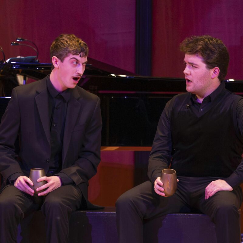 Two men wearing black suits sit next to each other, each holding a metallic cup. Both have expressive facial expressions, as if they are in the midst of a dramatic conversation or performance. A grand piano is visible in the background.