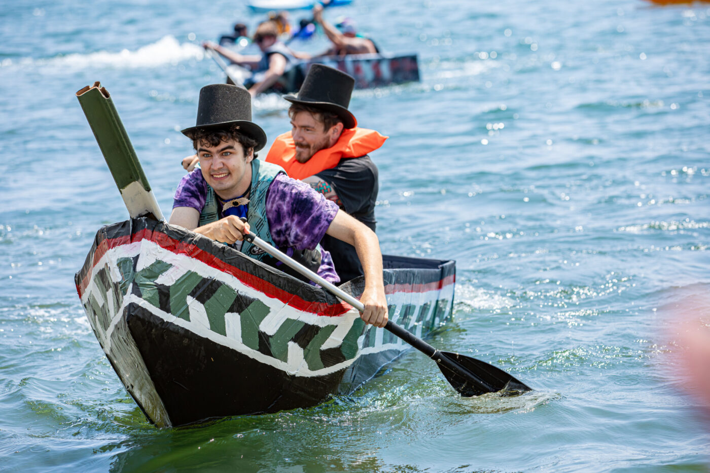 During Salem Heritage Days, two people in top hats and colorful shirts paddle a handmade boat, painted for the 400 Years celebration in 2026. Others join them on the water, enjoying the sunny day as smiles and laughter fill the festive atmosphere.