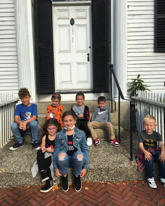 Six children sit on stone steps in front of a white building with a closed white door and black shutters. The kids are smiling, wearing casual clothes, and some hold small items. The steps are bordered by metal railings and fallen leaves are visible.