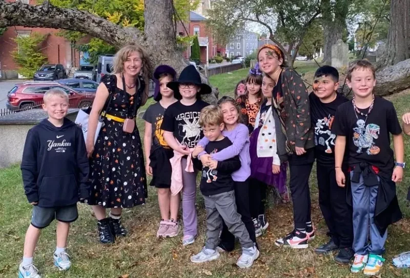 A group of children and one adult stand outdoors near a large tree. Many are dressed in black or Halloween-themed clothing. They are smiling and posing for the photo, with parked cars and brick buildings visible in the background.