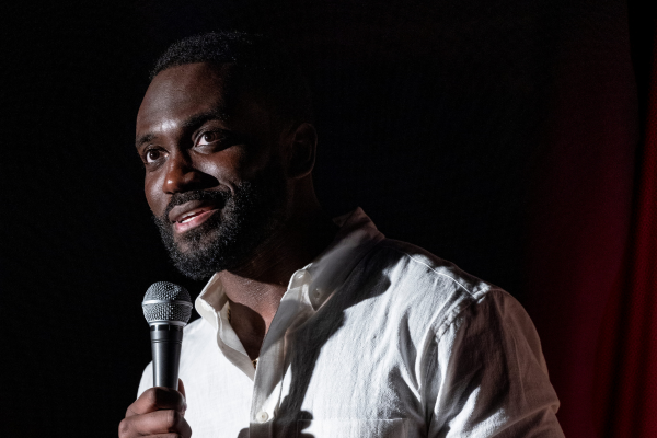 A man with a beard and mustache, wearing a white shirt, holds a microphone and speaks on stage. He is lit by a spotlight, while the background is dark.