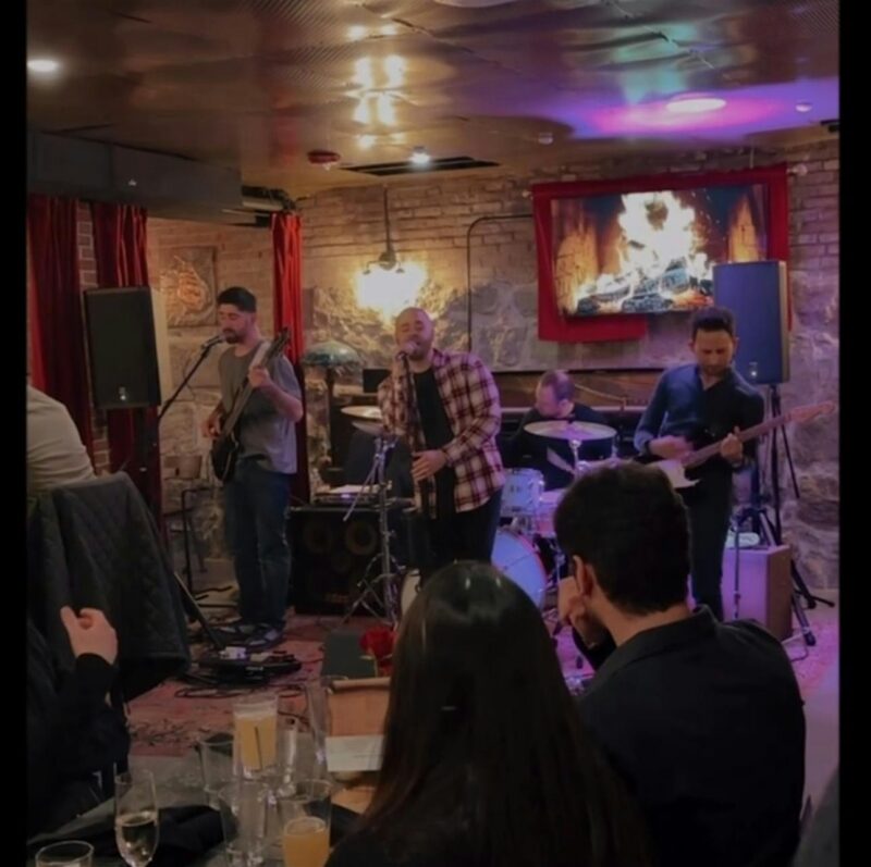 A four-member band performs in a cozy restaurant with exposed brick walls. People are seated at tables in the foreground. A digital fireplace image is displayed on a TV behind the band. Musical instruments include guitar, drums, and a microphone.