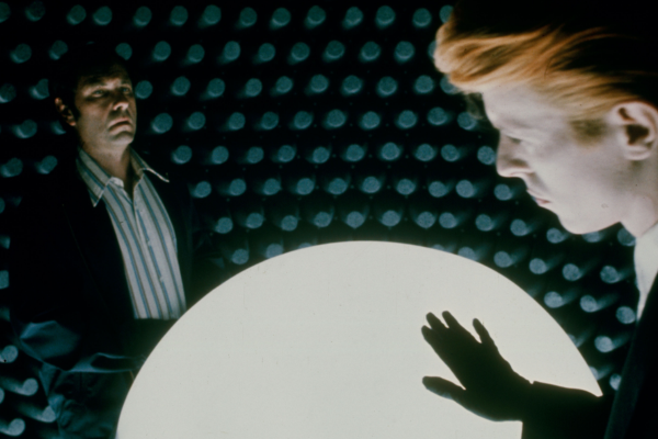 Two people stand in a room with soundproof foam walls. One person, in the foreground with orange hair, touches a glowing white sphere. The other, in the background, looks on seriously.
