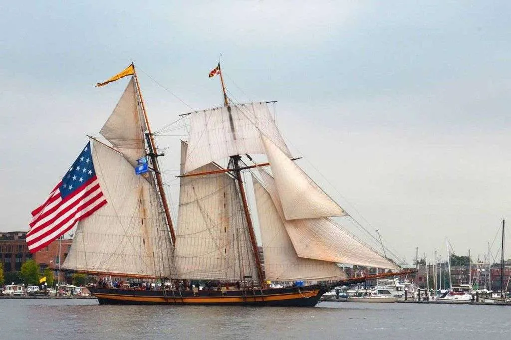 A tall wooden sailing ship with large sails is docked in a harbor for Salem Heritage Days. The historic American flag waves at the stern, with people on deck and other boats and buildings in the background under a cloudy sky.