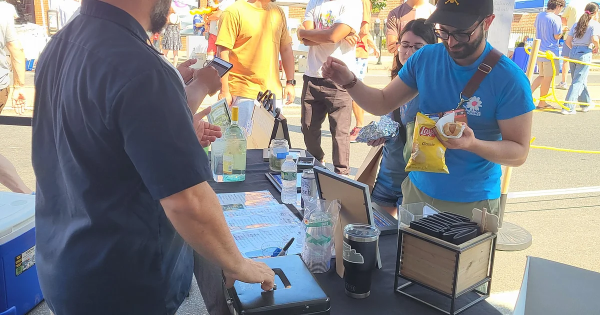 People stand at an outdoor booth covered with drinks and utensils. One person holds Lay's chips and a hot dog, while another interacts with a cash box. Other people and festival activities are visible in the background.