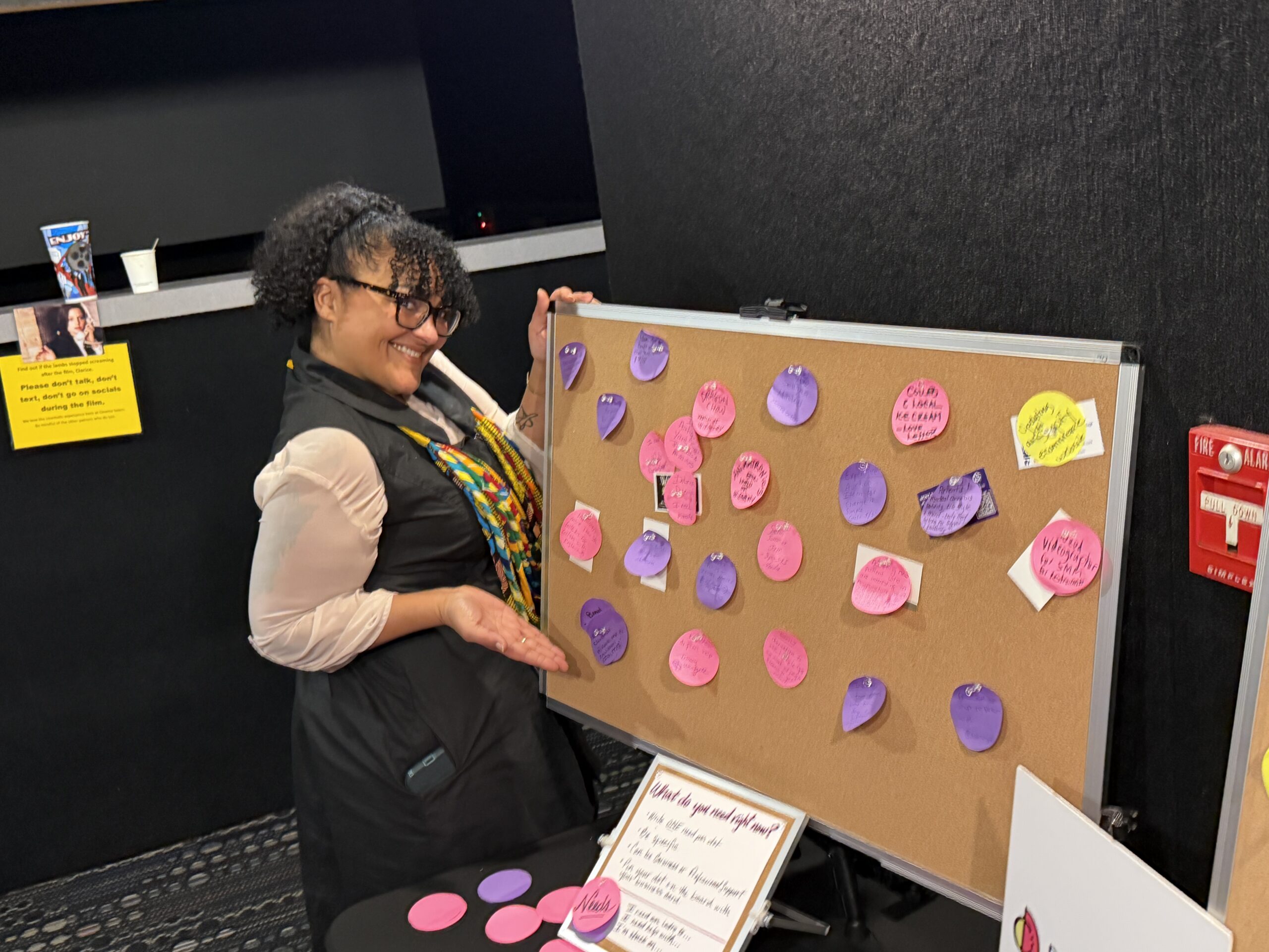 A person stands next to a corkboard displaying colorful paper circles with handwritten notes for Minority Mental Health Month. Smiling, they gesture toward the board. A small table with more circles and a sign is in front.