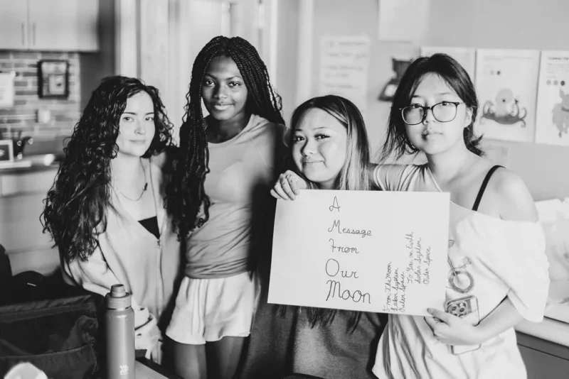 Four young women stand indoors at a Salon Show. One holds a handwritten sign reading, “A message from our mom.” They pose together in black and white; various objects and posters decorate the background.