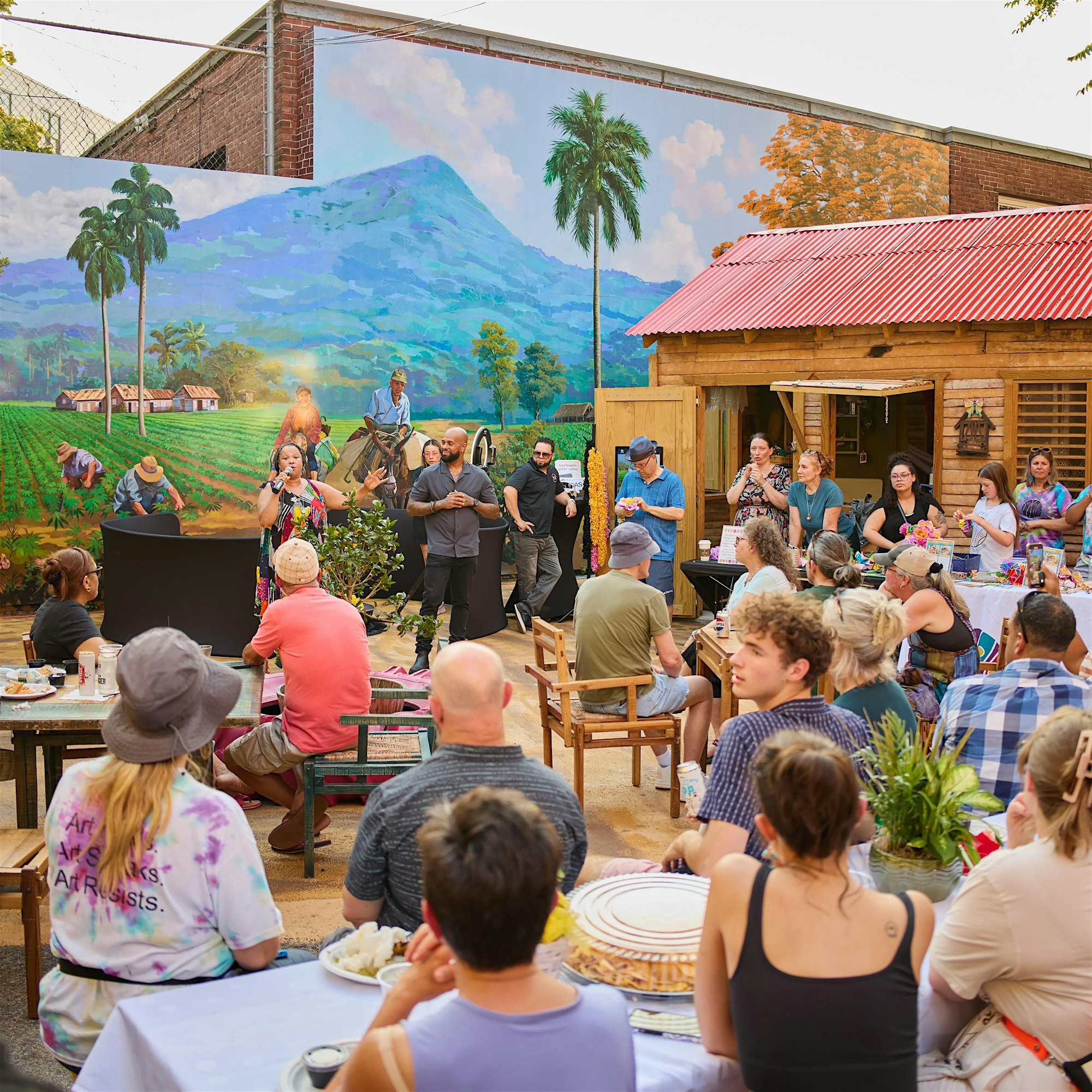 A group of people sit at tables outdoors, listening to a speaker at a podium. Behind them is a mural of a farming scene with mountains, palm trees, and workers. Some attendees are eating or talking, and a wooden building is visible in the background.