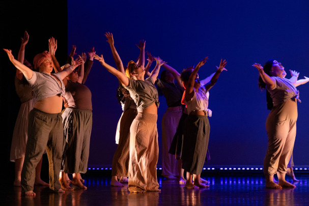 A group of barefoot dancers on stage, wearing loose, neutral-colored clothing, stand with their arms raised and heads tilted upward against a blue-lit background.