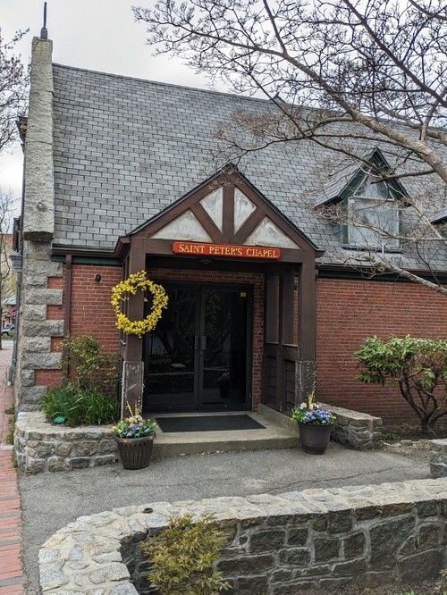 A small brick chapel with a steep gray shingle roof, wooden entrance, and a sign reading “Saint Peter’s Chapel.” There are planters with flowers and a yellow wreath decorating the doorway. Leafless branches are visible in the foreground.