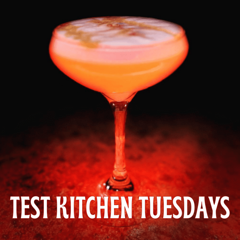 A cocktail glass filled with a pink-orange drink sits on a reddish surface. The text “TEST KITCHEN TUESDAYS” is written in large white letters at the bottom of the image.