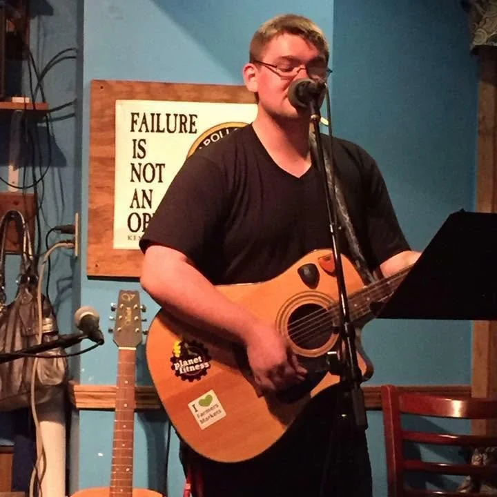 A person wearing glasses plays an acoustic guitar and sings into a microphone on stage. A sign reading “Failure is not an option” is on the blue wall behind them. Another guitar, a bag, and a chair are nearby.