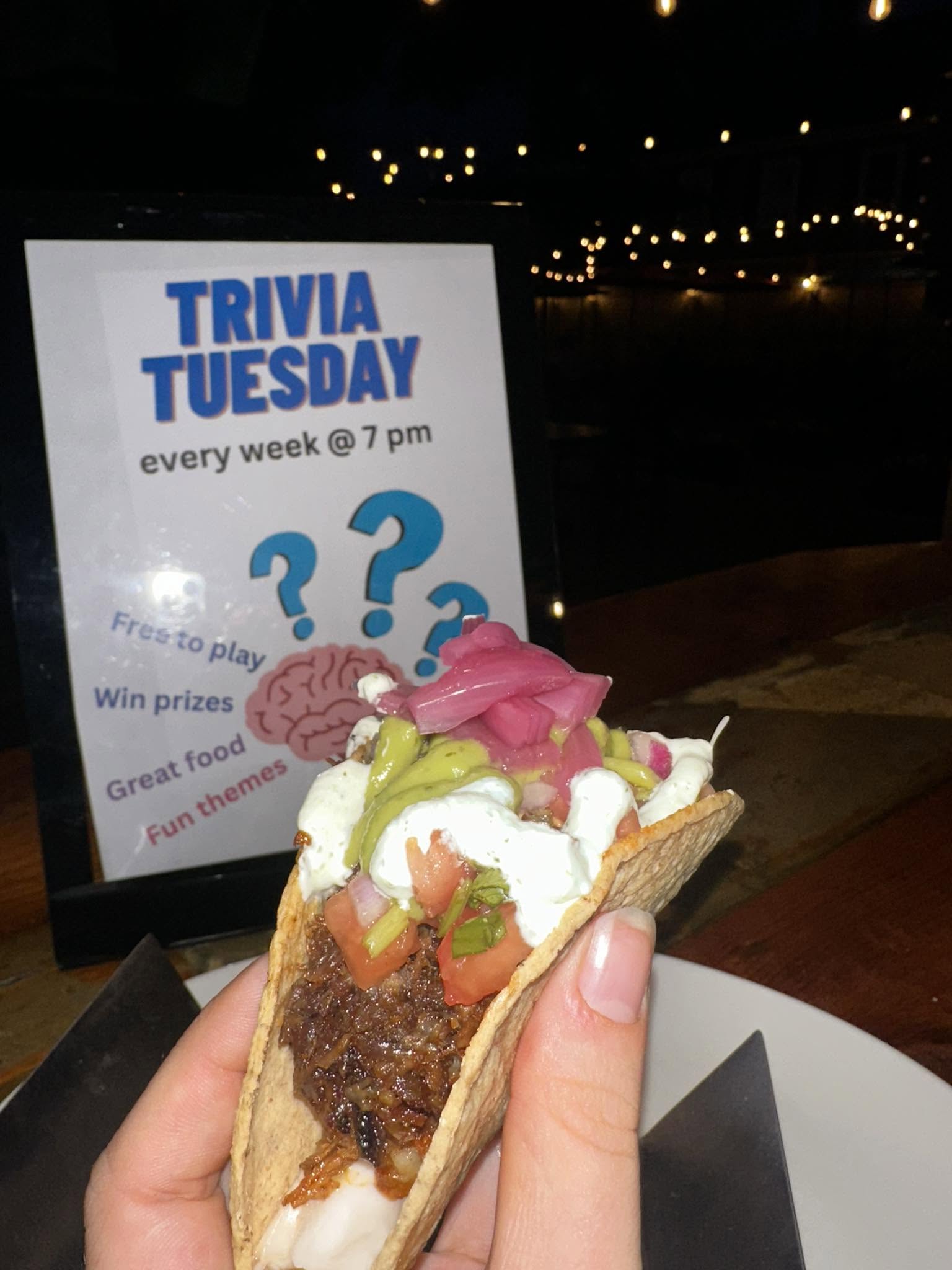 A hand holds a taco topped with vegetables and sauce in front of a sign for “Trivia Tuesday,” promoting weekly trivia at 7 pm with food, prizes, and fun themes. String lights are visible in the dimly lit background.