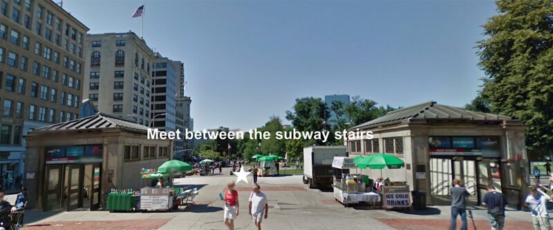 Two subway entrances stand across from each other in a public square with green umbrellas, food stands, and people walking. The words “Meet between the subway stairs” appear in the center of the image. Buildings and trees are in the background.
