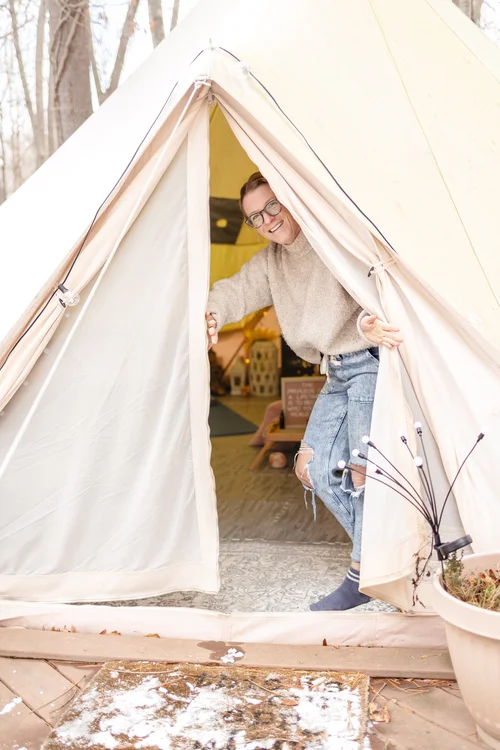 A person wearing glasses, a beige sweater, and ripped jeans smiles at the entrance of a beige tent in a wooded outdoor setting—perfect for a Sister Circle retreat. A doormat with snow and a potted plant sit by the tent’s entrance.